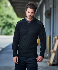 A man looking down with his arms by his sides wearing a black NATO security jumper and black cargo trousers.