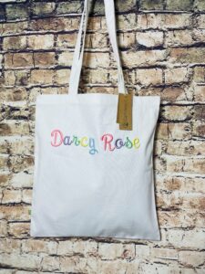 Frontal view of a natural-coloured tote bag against a brick background. On the front of the bag is text embroidered in 3D with letters of alternating pastel colours. The text reads: "Darcy Rose" in a cursive font.