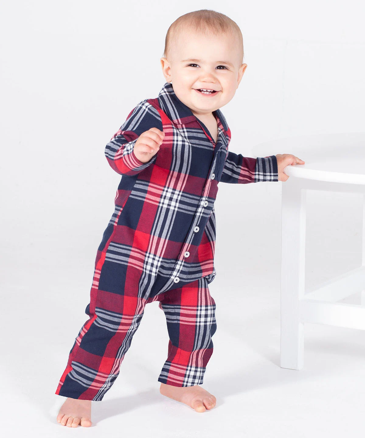 A smiling infant stood leaning against a white chair wearing the tartan all-in-one