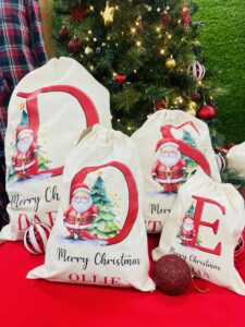 Natural coloured sacks of varying sizes with letters printed in red decorated with christmas trees and Father Christmas. Below, in cursive writing, reads "Merry Christmas". Below that, a different name on each sack matching the respective letter on each sack