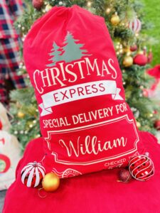 A red sack printed to the front with golden text that reads: "Christmas express special delivery for William checked twice". Above "Christmas" appears two green Christmas trees. "Express" is in red atop a white banner. "William" is within the outline of a frame. Below both "checked" and "twice" appears a ticked checkbox - one for each word. At the bottom left of the print is a a golden deer within two golden concentric circles. In the gap between the two concentric circles reads text: "north pole santa mail". To the left of and slightly overlapping the concentric circles and deer are four wavey golden lines imitating those found on a marked stamp.