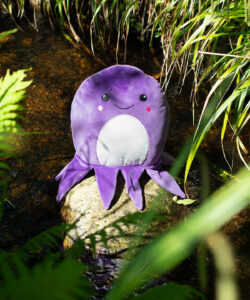 Frontal view fo the purple octopus squidgy outside on top of a stone in the middle of a stream with water running either side of it. Long green leaves droop down into the stream.