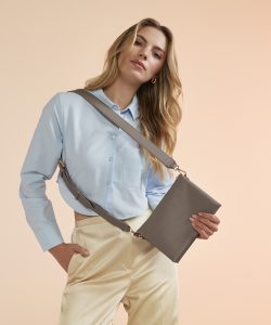 A woman with blond dyed hair wearing a blue cropped blouse and cream trousers holding her hand the Athena cross-body bag in taupe whilst it is hung over her shoulder