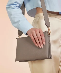 A woman's hand with painted nails holding the Athena cross-body bag in taupe whilst it's hung over her sholder.