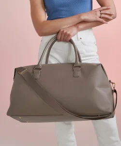 A woman in a blue top and white trousers holding a taupe Iris weekender bag in front of her by its top handles