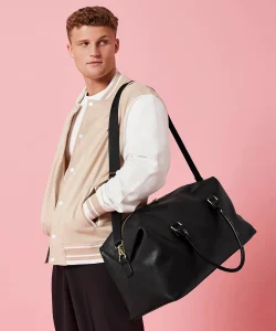 A man with brown hair wearing a cream varsity jacket with white sleeves and a black Iris weekender bag with golden metal meatures hung over his shoulder by its shoulder strap