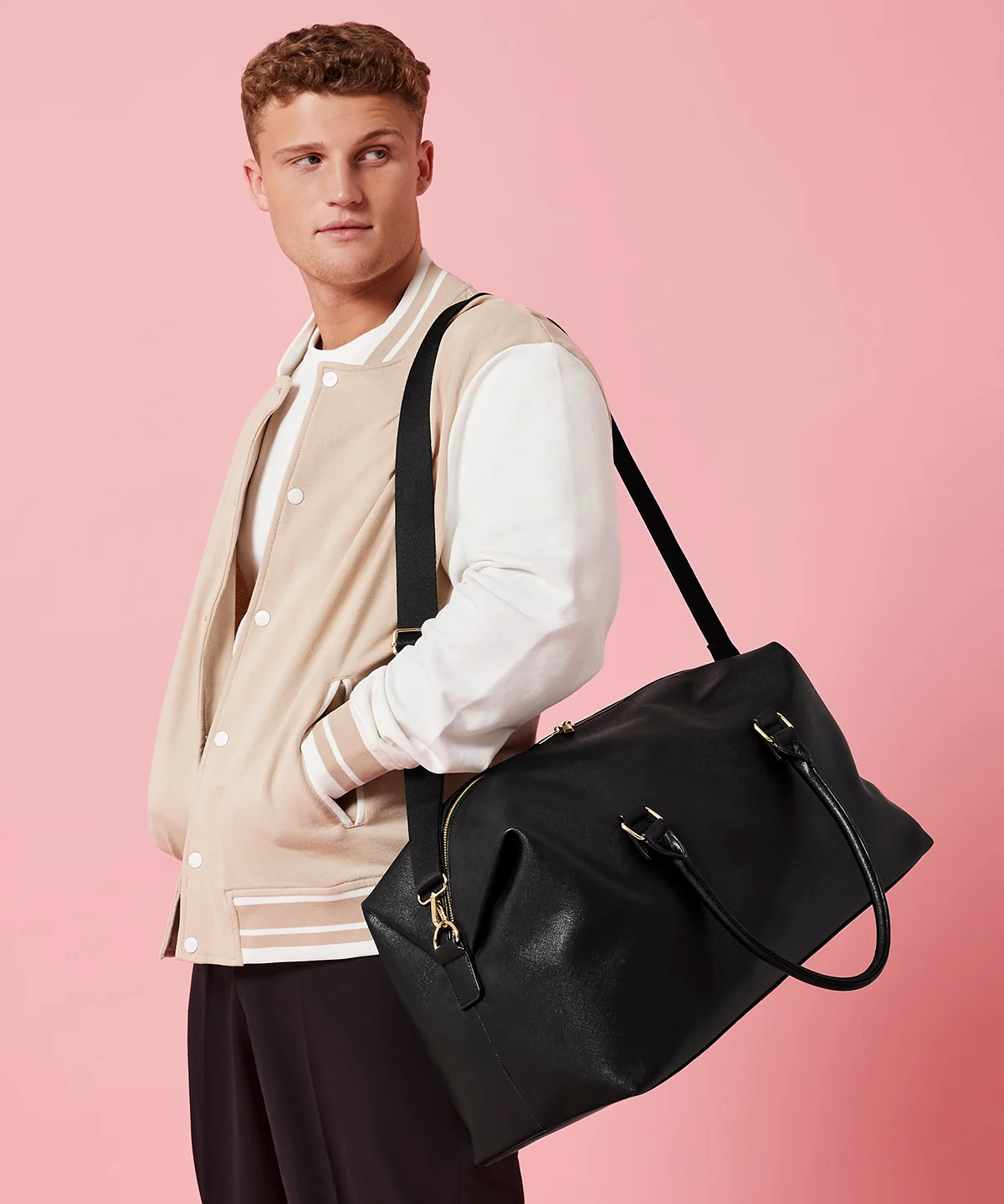 A man with brown hair wearing a cream varsity jacket with white sleeves and a black Iris weekender bag with golden metal meatures hung over his shoulder by its shoulder strap