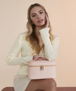 A woman with brown hair sat smiling wearing a cream-coloured cardigan and white satin mini skirt in front of a beige background holding the Aphrodite vanity case in soft pink resting on her knee.