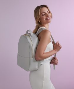 A woman with dyed blonde hair smiling, wearing a white dress with the Penelope backpack in soft grey hung over her shoulder with one of the back straps