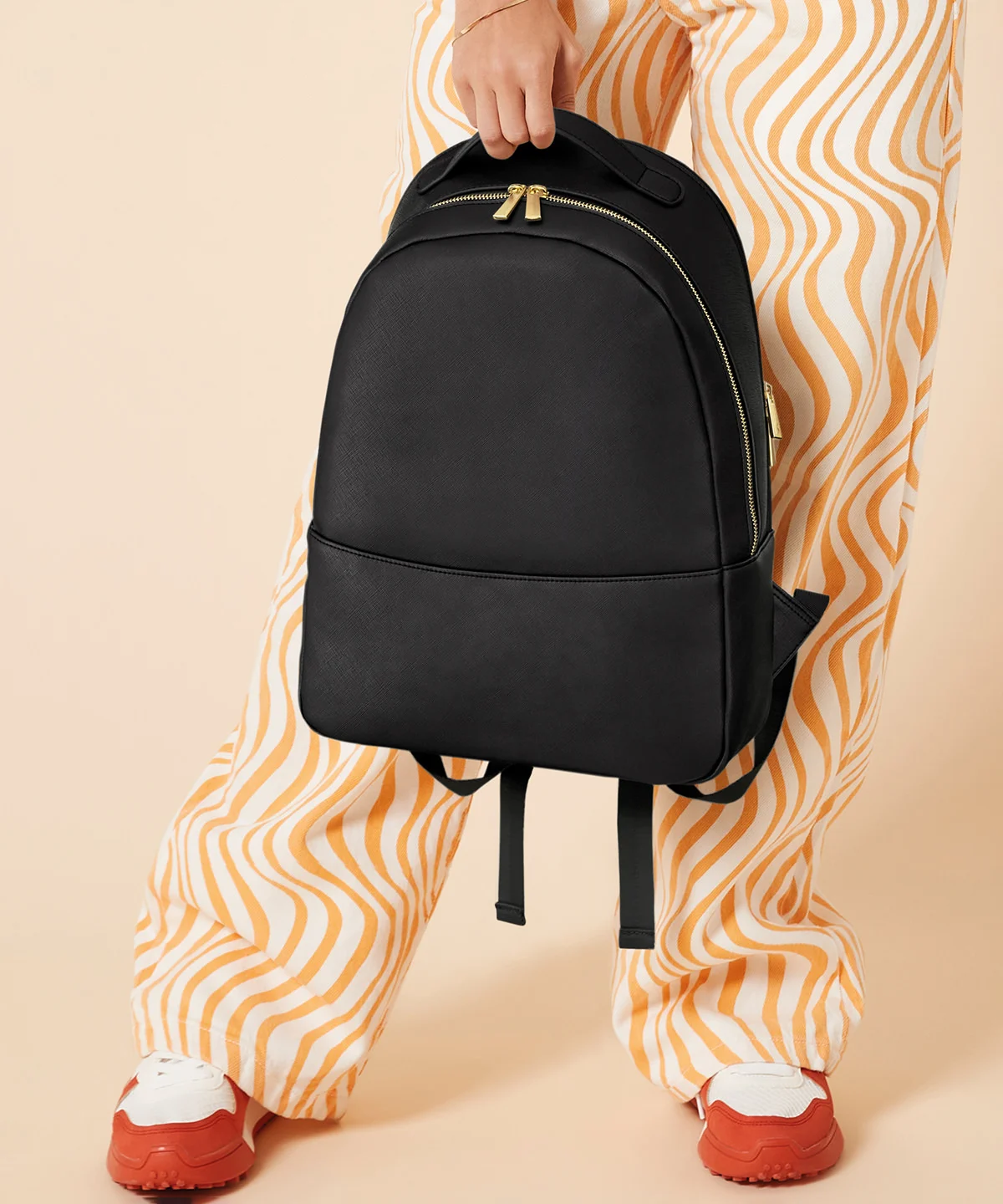 A woman in orange and cream wavey trousers holding the Penelope backpack in black in front of her with one hand by the grab handle on top