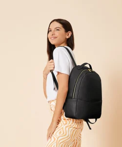 A woman with dyed blonde hair wearing a white dress with the Penelope backpack in soft grey hung over her shoulder with one of the back straps