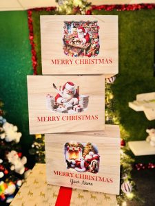 Frontal view of three wooden boxes sat atop a wrapped Christmas present in front a Christmas Tree. On the first box is a picture of Santa in his workshop surrounded by colourful toys using a sewing machine. On the second box is a printed picture depicting Santa writing a letter with a quill and surrounded by piles of already complete letters. On the third box is an image printed depicting Santa sat in an arm chair with his feet up. Behind him is a fireplace with three red stokcings hanging up. Next to the fireplace is a well decorated Christmas tree.