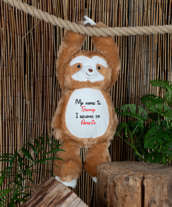 The large personalised sloth teddy hanging from a rope by its hands connected together with hook and loop fastening. Behind the teddy is a bamboo background with tropical indoor plants.