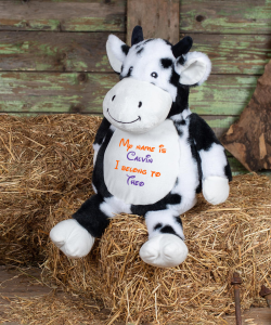 A cow teddy sat atop a hay bale in front of a wooden board backdrop. On the belly of the cow is sample personalisation that reads: "My name is Calvin I belong to Theo". The text is in orange and purple.