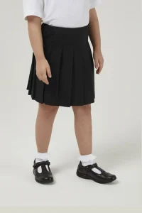 A young girl wearing the junior black stitched-down premium pleated skirt with a tucked-in white blouse and white socks with school shoes.