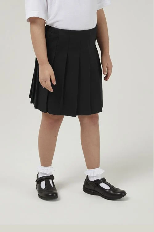 A young girl wearing the junior black stitched-down premium pleated skirt with a tucked-in white blouse and white socks with school shoes.