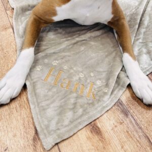 A closeup view of the name "Hank" embroidered on a beige dog blanket with a boxer dog's paws either side.