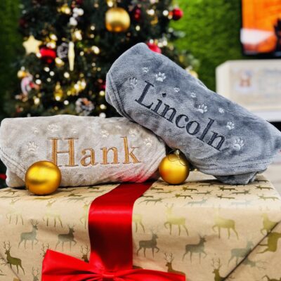 Two personalised dog blankets rolled up on top of a present with baubles in front. The blankets are beige and grey with names "Hank" and "Lincoln" embroidered respectively and paw prints around them.